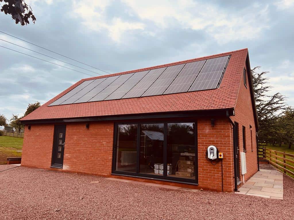 A modern red brick outbuilding with a large array of solar panels mounted on its red tiled pitched roof, featuring a wall-mounted EV charging point.