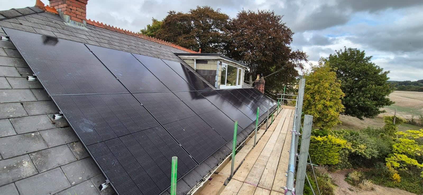 A newly installed array of sleek, black solar panels on a grey slate tiled roof next to a dormer window, with scaffolding running along the roof edge.