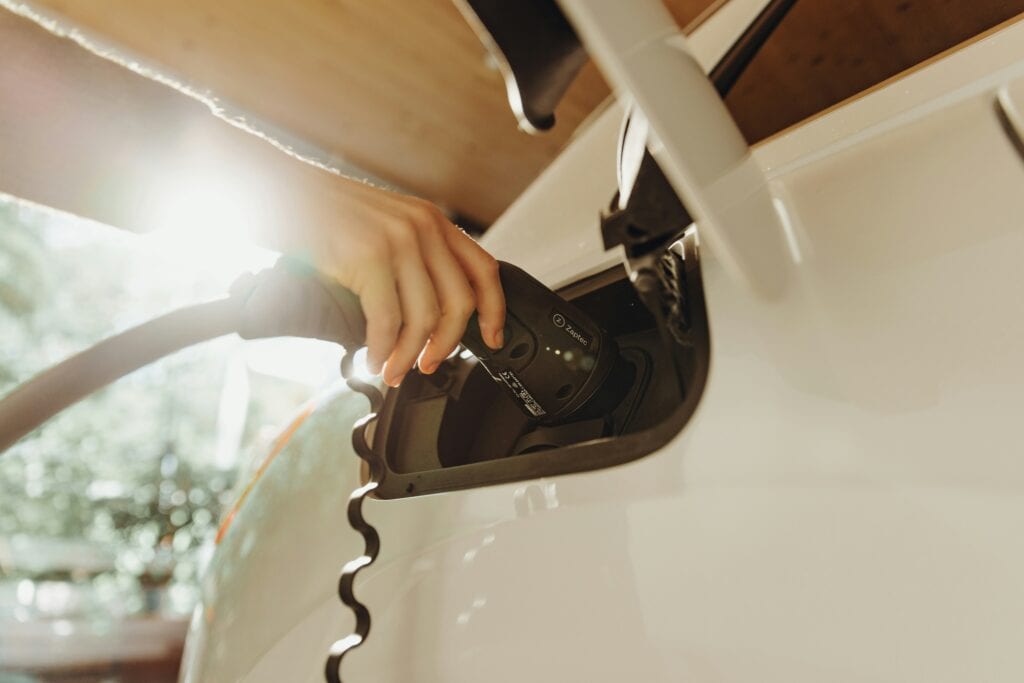 Close-up of a person's hand plugging a black EV charging cable into the open charging port of a white electric car, backlit by bright, warm sunlight.